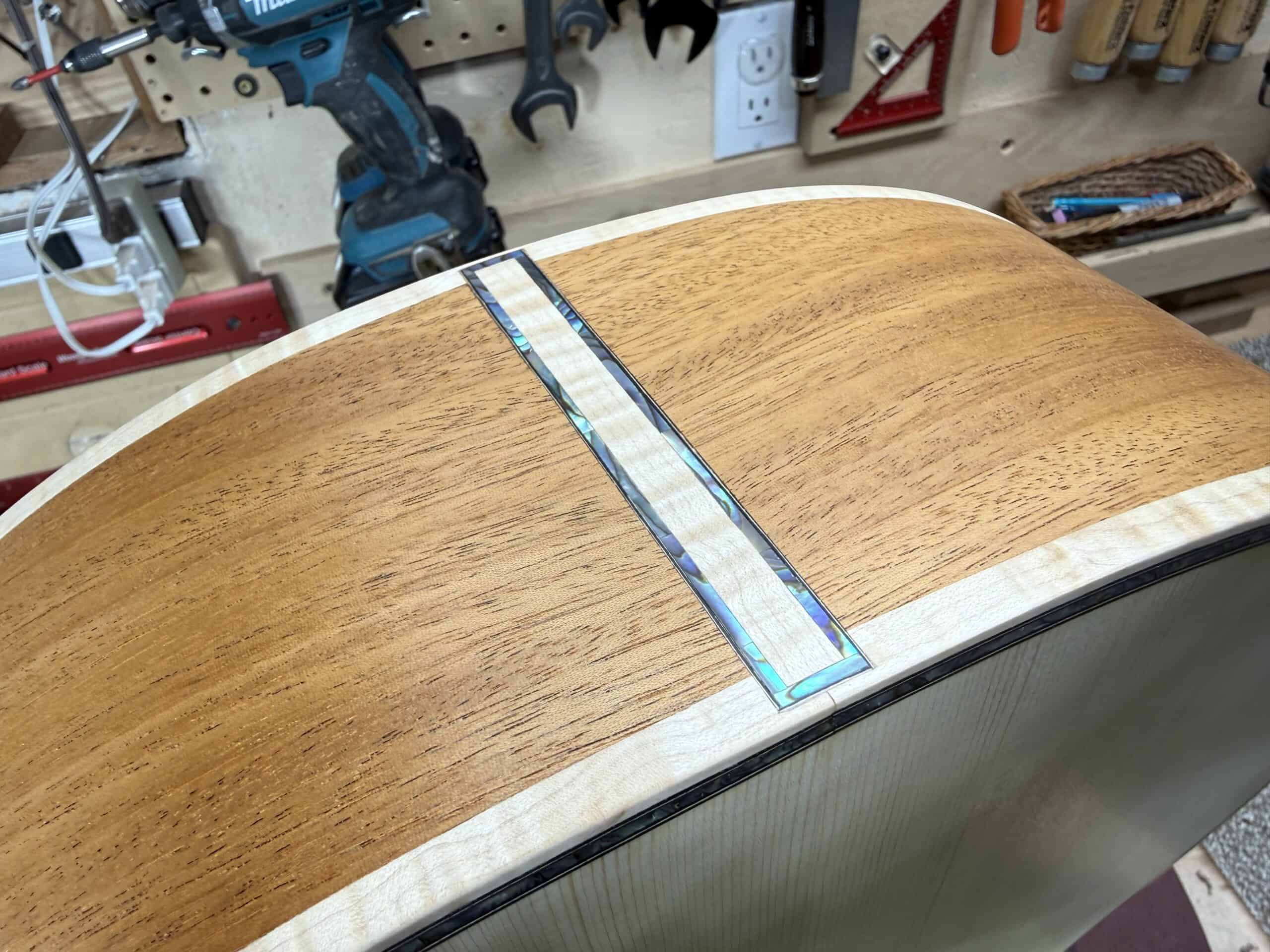 Custom acoustic guitar body in workshop with woodworking tools and workbench background.